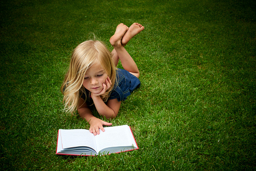 Cute Little Blond Girl Reading Book Outside on Green Grass Lawn.