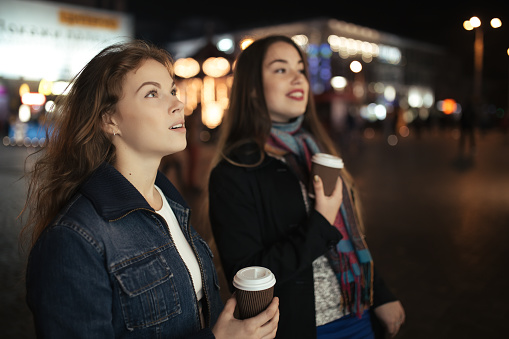 Two women friends walking in city street and drinking coffee at night