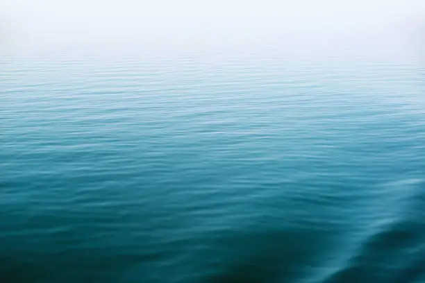 Small ripples and soft waves on the surface of a calm, deep lake. Horizon is hidden in fog.