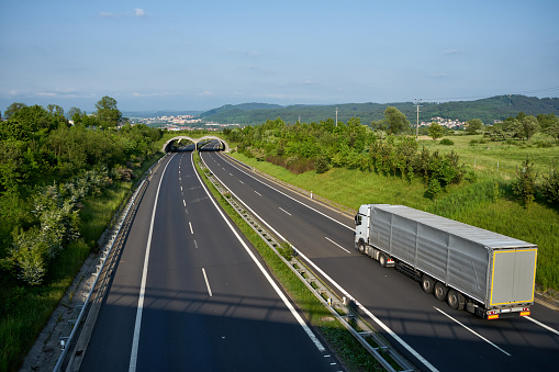 Asphalt highway with the ecoduct with driving white truck, city and forested mountains in the background. View from above.