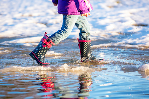 Waist down side view of a young girl walking through a large puddle of melted snow on an early springtime evening. The bottom of a purple jacket is visible, as well as light denim blue jeans, and navy blue with white polka dots and pink trim rubber boots.