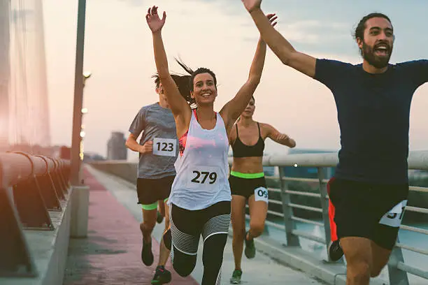 Runners running marathon in the city. They are running over the bridge at sunset. Wearing numbers on their sport clothes. Smiling. Running through the finish with arms raised.