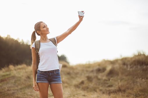 Beautiful young blonde hiker girl is taking selfie in nature on warm sunny day