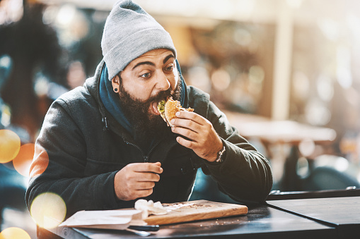 Closeup of bearded hipster guy gorging on a burger like if nobody's watching. He's sitting outdoors on a cold sunny day. He's wearing a dark jacket, gray cap and has long brown beard.