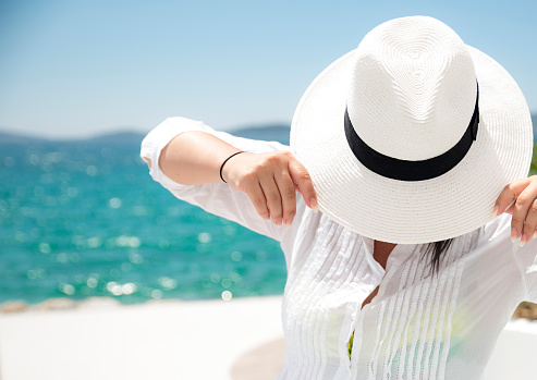 Woman in white shirt standing by the sea and holding a white hat over her face with head down. Photo of waist up.