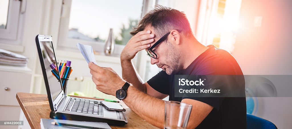 Man reading letter and felling worried Man sitting in home office, reading letter and felling worried. With one hand holding letter and with other hes forehead Financial Bill Stock Photo