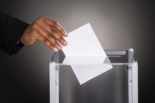 Close-up Of A Businessperson Hand Inserting Ballot In Glass Box Against Gray Background
