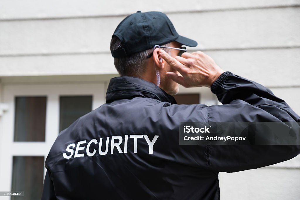 Security Guard Listening To Earpiece Against Building Rear view of mature security guard listening to earpiece against building Security Guard Stock Photo