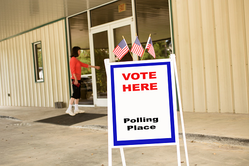 'Vote Here, Polling Place' sign outside of a local, public polling location, which is a public church in USA.  American flags top the sign.  One woman enters the building.  The USA elections are held in November each year.  Sign created by photographer.