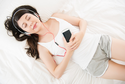 asian woman enjoy music by smart phone with headphones lying on bed. home indoors background. high angle view