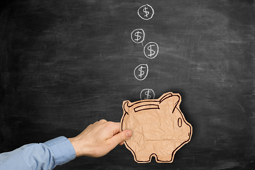 businessman holding a piece of brown crumpled paper cut in form of a piggy bank  over hand drawn falling coins on blackboard