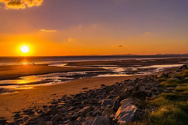 The sun setting over the sea from Barmouth Bay, Snowdonia National Park in Wales, UK. Mountains on the Horizon with rocky foreground and the sandy beach. 