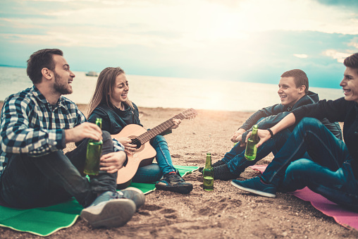 Four Smiling People with Acoustic Guitar Singing on Beach Camping. They are having summer fun on the sunrise.