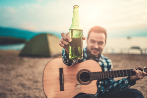 Young Guitar Player Camper on Beach with Raised Beer Bottle for Cheers. He is having summer fun on the sunrise.