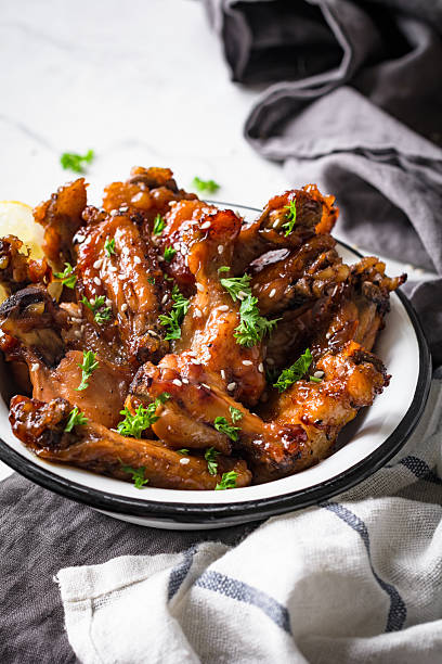 Chicken wings barbecue Teriyaki chicken wings in white bowl garnished with sesame seeds, chopped parsley and lemon wedge on marble table. Side view, toned, vertical sticky sesame chicken sauces stock pictures, royalty-free photos & images