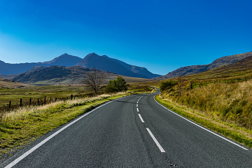 A winding mountain road in Snowdonia National Park, Wales, UK. Mountain range in the background with the empty road winding into the distance on a clear, sunny, summers day. 