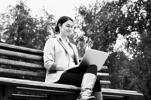 Relaxed woman sitting on a bench in park, working on a laptop. Black and white shot.