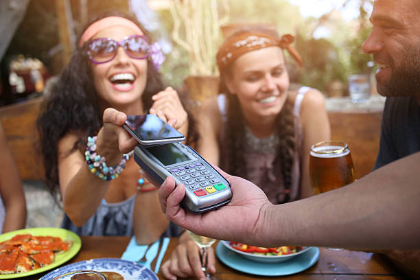 Cheerful young woman using smart phone for mobile payment Young people having fun in restaurant. Smiling young woman using smart phone for contactless payment. All with casual clothes. Food and drinks on table in front of them. On foreground human hand holding mobile pos terminal. Focus on foreground. friends in bar with phones stock pictures, royalty-free photos & images