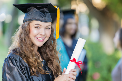 Beautiful Caucasian college girl smiles proudly while holding her diploma. She has long curly brown hair and is wearing a black cap and gown.