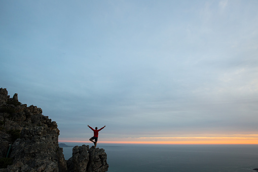 A male stands on one leg doing a yoga move on a high mountain peak at sunset