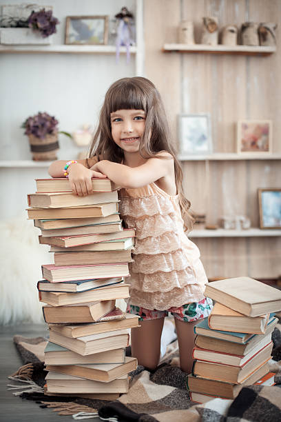 istock little cheerful girl with stack of books in library; 614702186