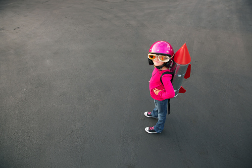 A young girl standing on a road, wearing jeans, a red shirt, and black shoes. She is equipped with oversized goggles and a large pink helmet.  He has a homemade rocket strapped to her back, with a silver body and red fins and top.  The girl is looking at the camera