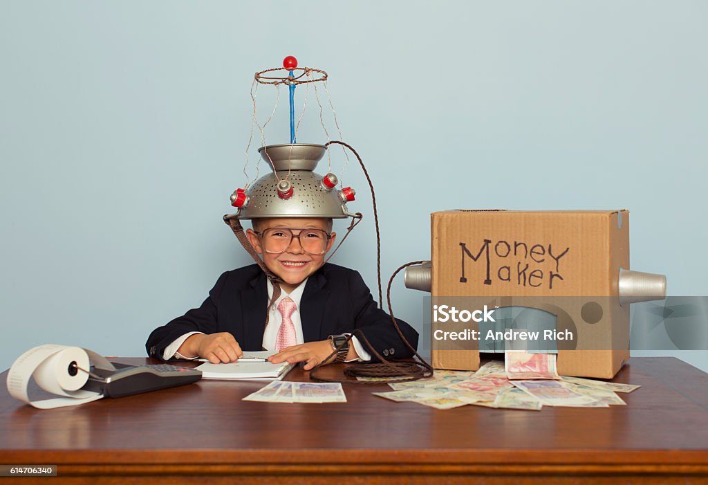Young Businessman Makes Money with Homemade Money Machine A young boy businessman sitting at a desk records the amount of British Pound Sterling his homemade money machine makes. He is dressed in a suit and pink tie, glasses, and a mind reading helmet on his head. Let's make some money with clever business. Interest Rate Stock Photo