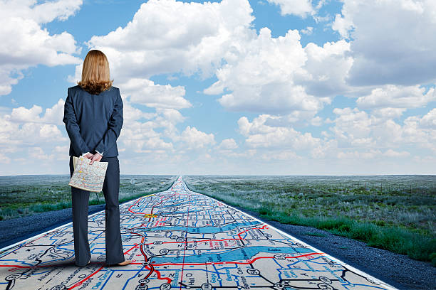 Businesswoman Stands On Long Road With Road Map Painted On It A businesswoman stands while holding a road map in her hands as she looks down a long, straight, rural road that has the same road map painted on it. road map stock pictures, royalty-free photos & images