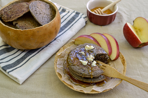 Rustic, vintage meal of buckwheat pancakes in plate and bowl made of gourds, on flower sac tablecloth, accompanied by apple slices and maple syrup, viewed from above