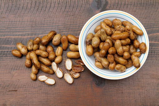 Bowl of boiled peanuts, a deep south regional food. Very salty, mushy and addictive. Overhead shot against a dark wood background. A few broken open peanuts are with other whole peanuts on the table next to the bowl.