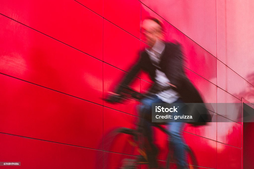 Handsome man riding bicycle beside the red wall Blurred image of a handsome man, on the way to work, riding bicycle beside the red wall. The man is casually dressed and wears eyeglasses and carries black briefcase hung on shoulder. Motion concept, motion blur, copy space has been left. Cycling Stock Photo