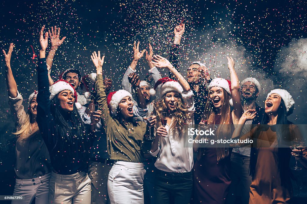 Celebrating New Year together. Group of beautiful young people in Santa hats throwing colorful confetti and looking happy Christmas Stock Photo