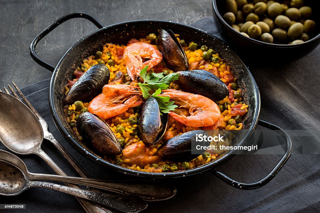 Paella on a table Paella in black pan with rice, shrimps, mussels, squid and meat, bowl with olives and vintage cutlery. Seafood paella, traditional spanish dish. Paella on rustic black wooden table. Selective focus Paella Stock Photo