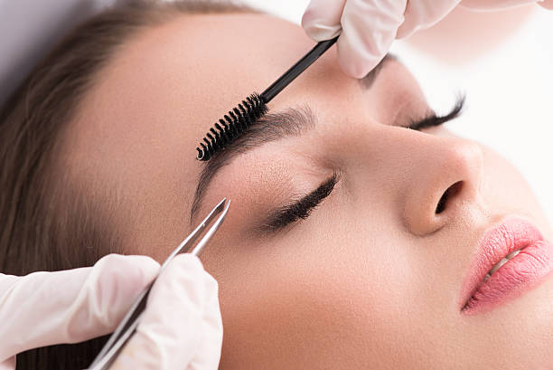 Young woman tweezing her eyebrows in beauty saloon Plucking for perfection. Cropped shot of woman in white gloves using tweezers on patient eyebrow at health spa isolated on white background eyebrow photos stock pictures, royalty-free photos & images