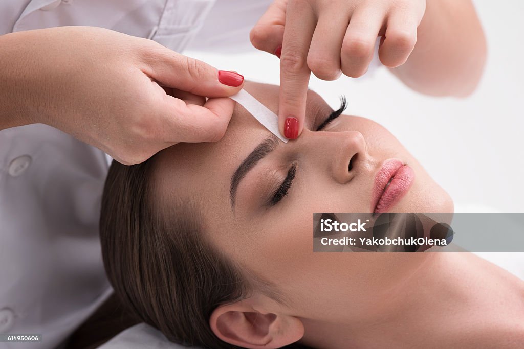 Young woman tweezing her eyebrows in beauty saloon Already perfect. Close up of hands waxing beautiful woman eyebrow, lying in beauty salon with closed eyes isolated on white background Waxing - Hair Removal Stock Photo