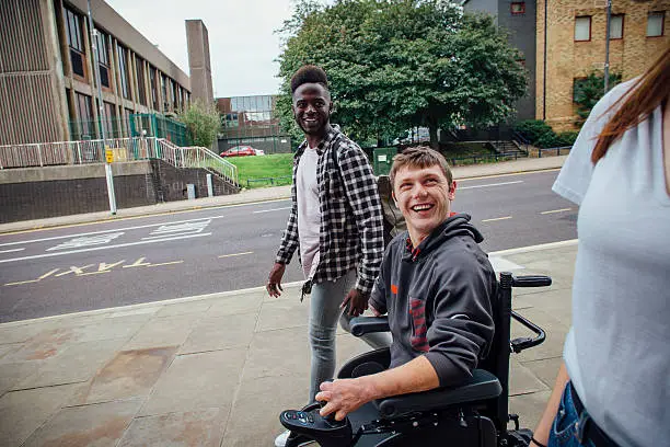 Three friends walking down the street together. One is quadriplegic and is in a wheelchair. Both guys are laughing and looking at the girl, who is only partially in shot.