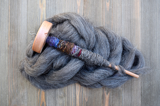 Wood drop spindle with natural sheep wool fiber twisted yarn wrapped around the dowel. The yarn is gray, blue, pink, and brown. The spindle is on top of a weathered wood table laying on top of a pile of natural gray wool roving fiber.