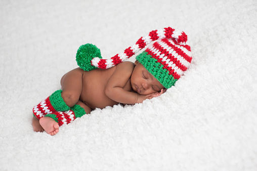 A one month old baby girl wearing a crocheted, red, white and green stocking cap and matching leg warmers. Photographed on a white, fluffy blanket.