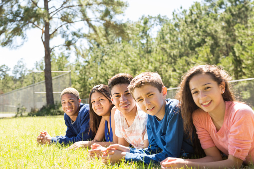 Multi-ethnic group of teenage friends hang out outside together at a local park or on a school campus. They are lying on their front in grassy field.  Nature in background.