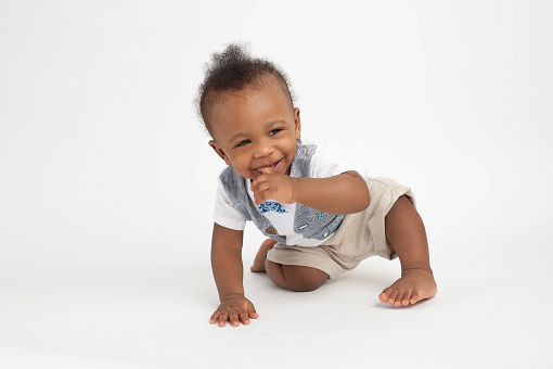 A smiling, nine month old, baby boy sitting on a white, seamless background with his finger in his mouth.