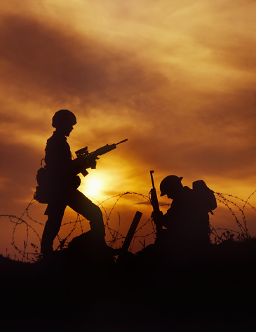 A silohouette photo of two soldiers surrounded by barbed wire stand in front of a dramatic evening sky. The men are darkened and made their faces to be unknown.