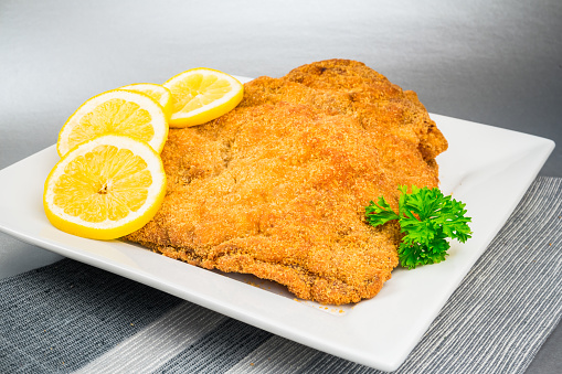 Photo of a portion of Wiener Schnitzel in a square white plate, lemon slices and green parsley as garnish.