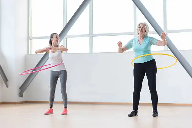 Developing body. Pleasant confident cheerful women standing in the gym hall and rotating hula hoops while having fitness training