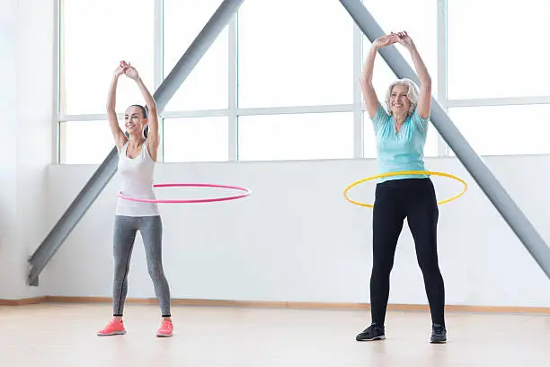 Making your waist thinner. Good looking energetic sporty women holding their hands up and smiling while rotating hula hoops
