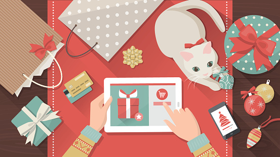 Woman purchasing Christmas gifts online using a tablet, her cat is playing with a bauble on the desk, holiday and celebrations banner