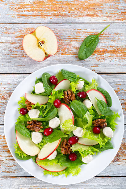 salad of apple, spinach, mini mozzarella balls, lettuce leaves, delicious autumn vitamins vegetarian salad: apple, spinach, mini mozzarella balls, lettuce, walnuts, cranberry on white dish  on table, vertical view from above green apple slice overhead stock pictures, royalty-free photos & images