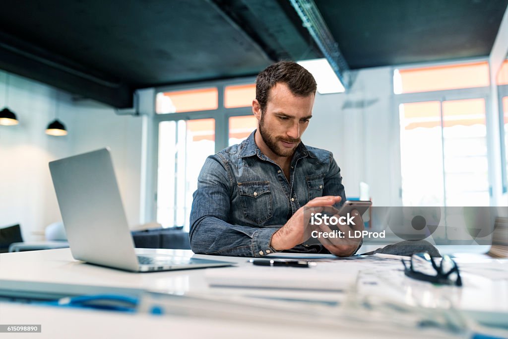 Man using a app mobile phone in modern office start-up. Young male in modern office start-up working on laptop. using smartphone. Sms, message, chat, texting Mobile Phone Stock Photo