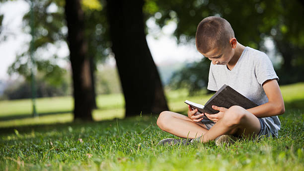 istock Cute schoolboy reading book in nature while sitting on grass 615240130