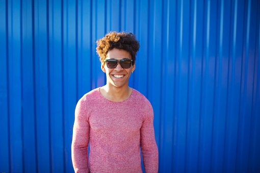young African American man over blue background wearing sunglasses. Cool teenager with stylish hairstyle smiling over color wall