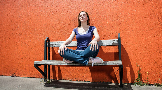 relaxing beautiful young woman enjoying meditating on a bench in a yoga position, breathing for spirituality and detox over an orange wall in the street, summertime outdoors